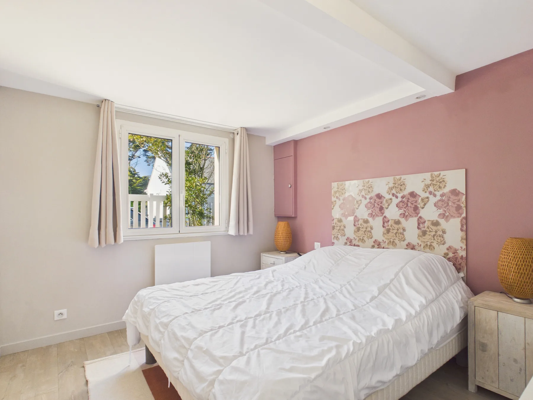 Dormitorio con una cama blanca, una ventana y decoración floral en la pared.