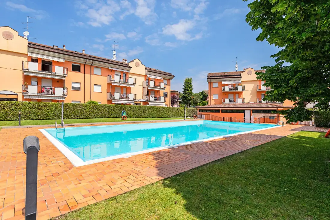 An outdoor swimming pool sits beside orange apartment buildings and a grassy lawn.