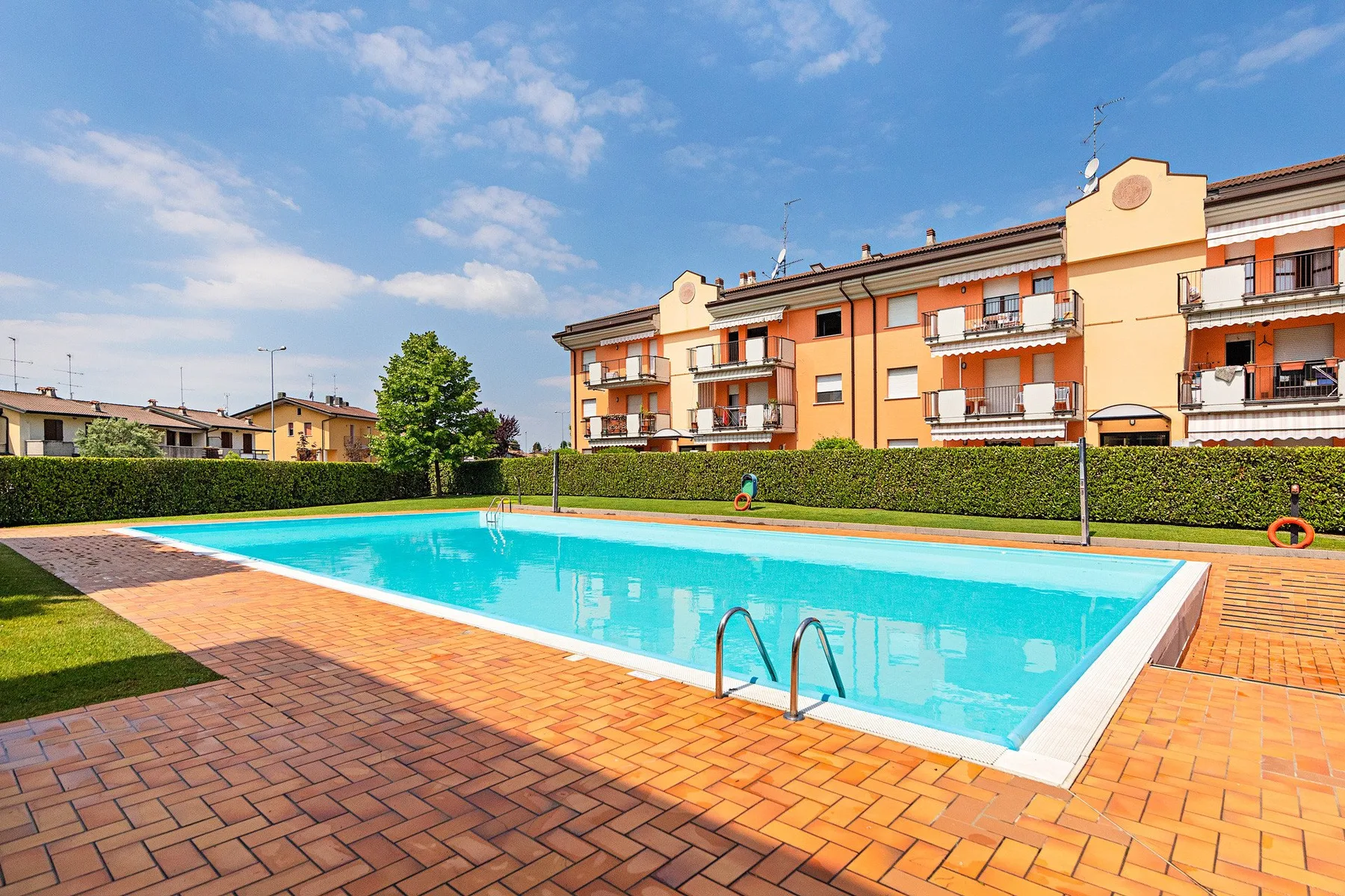 An outdoor swimming pool sits beside a brick-paved deck and apartment buildings.