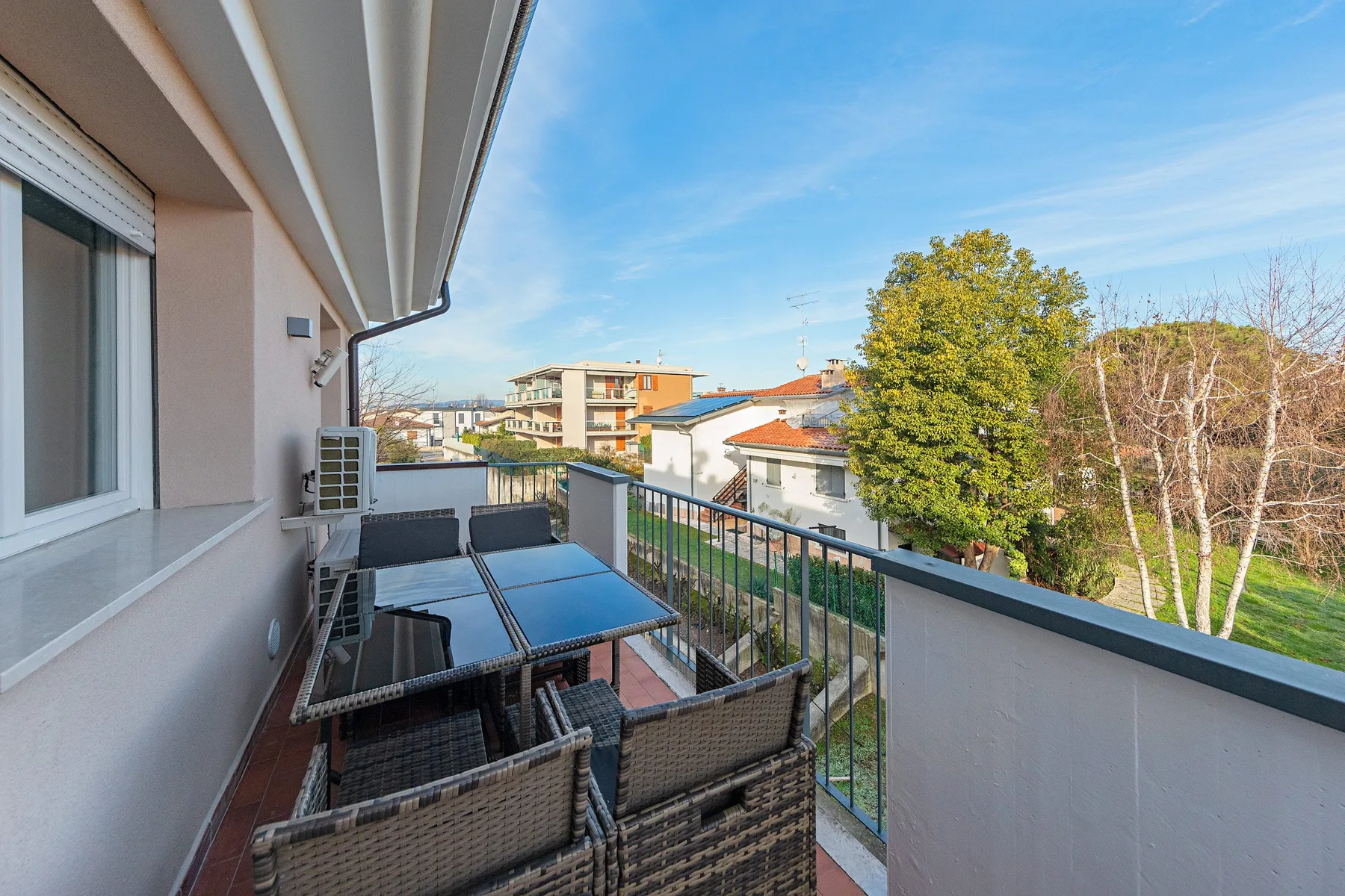 A balcony seating area with a table overlooks nearby buildings and trees.
