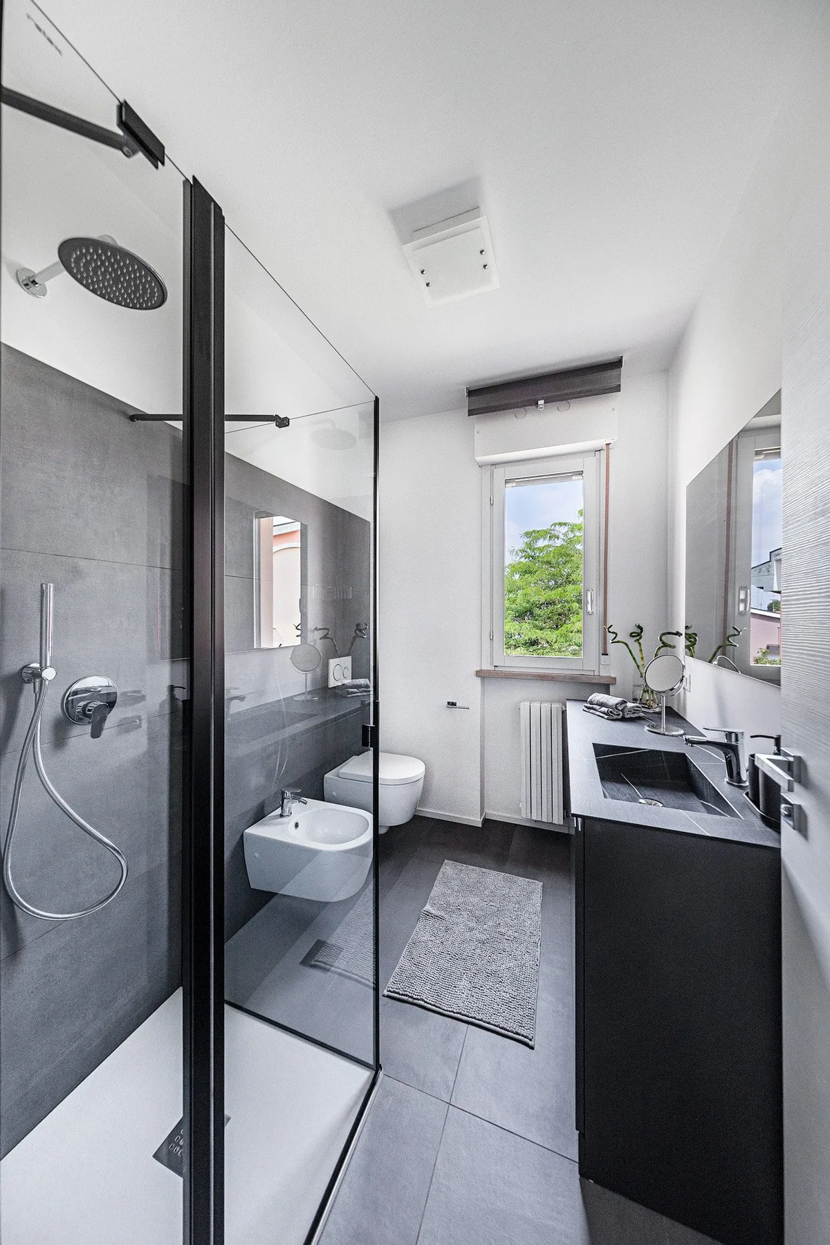 A modern bathroom with a glass shower, vanity, and window overlooking greenery.