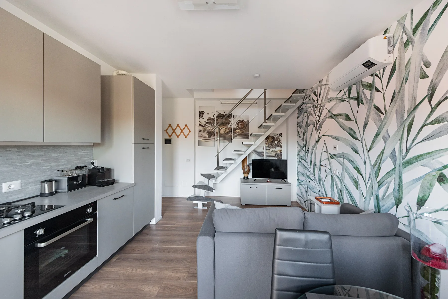 An open-plan living room and kitchen with a staircase and leafy wall mural.