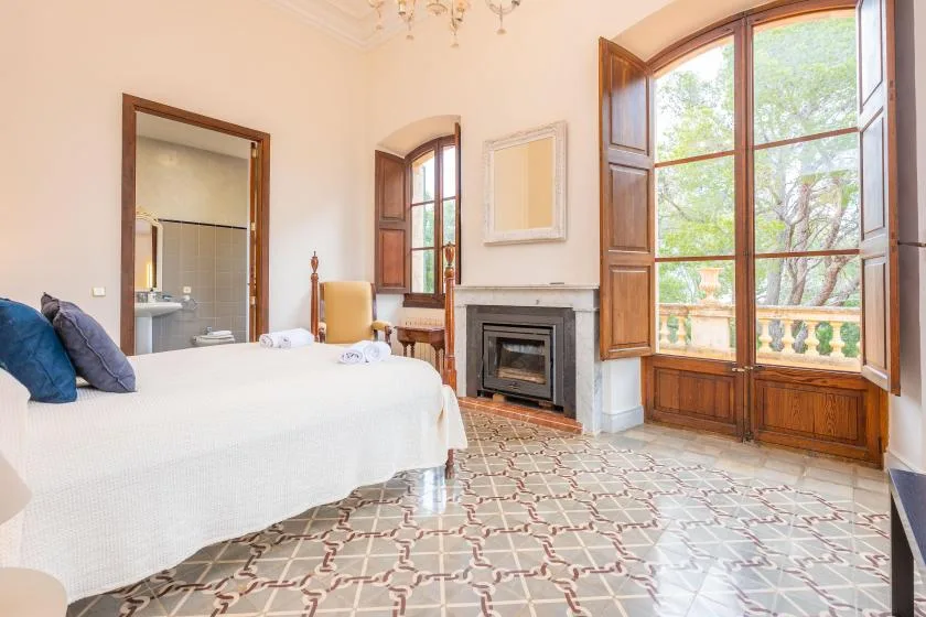 Un dormitorio luminoso con cama, chimenea y ventanas altas con vistas a la vegetación.