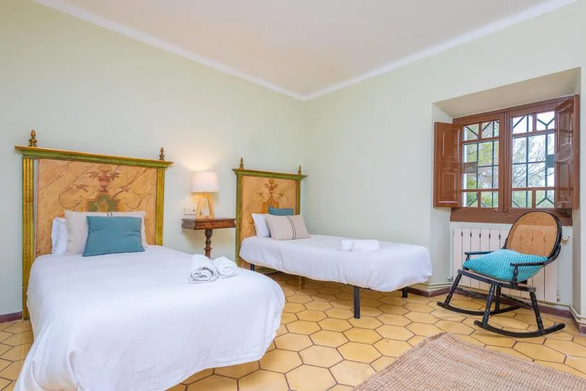 Un dormitorio con dos camas, una ventana con marco de madera y una mecedora.