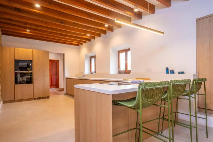 Una cocina luminosa con una isla grande y asientos verdes bajo vigas de madera.