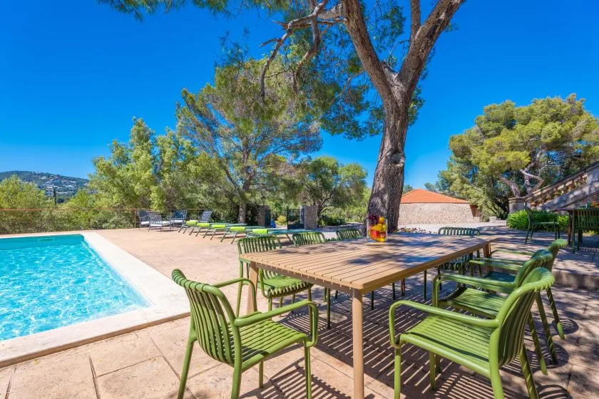An outdoor poolside patio has green seating around a wooden table beneath the trees.