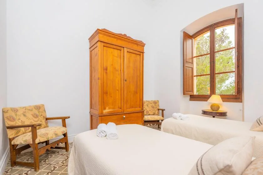 Un dormitorio luminoso con dos camas, asientos de madera y una ventana abierta.