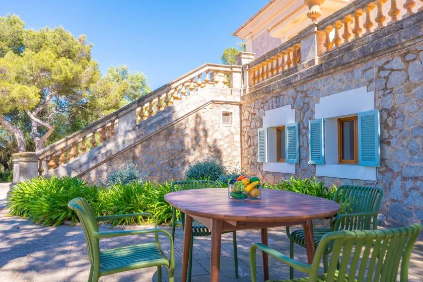 Una terraza de piedra tiene una mesa redonda, asientos verdes y una escalera junto al muro.