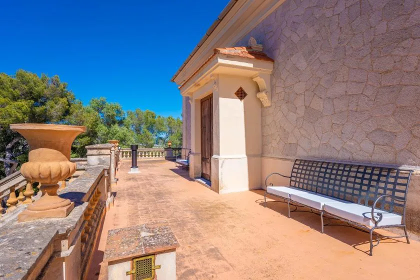 Una terraza de piedra soleada con un banco y árboles al otro lado de la baranda.