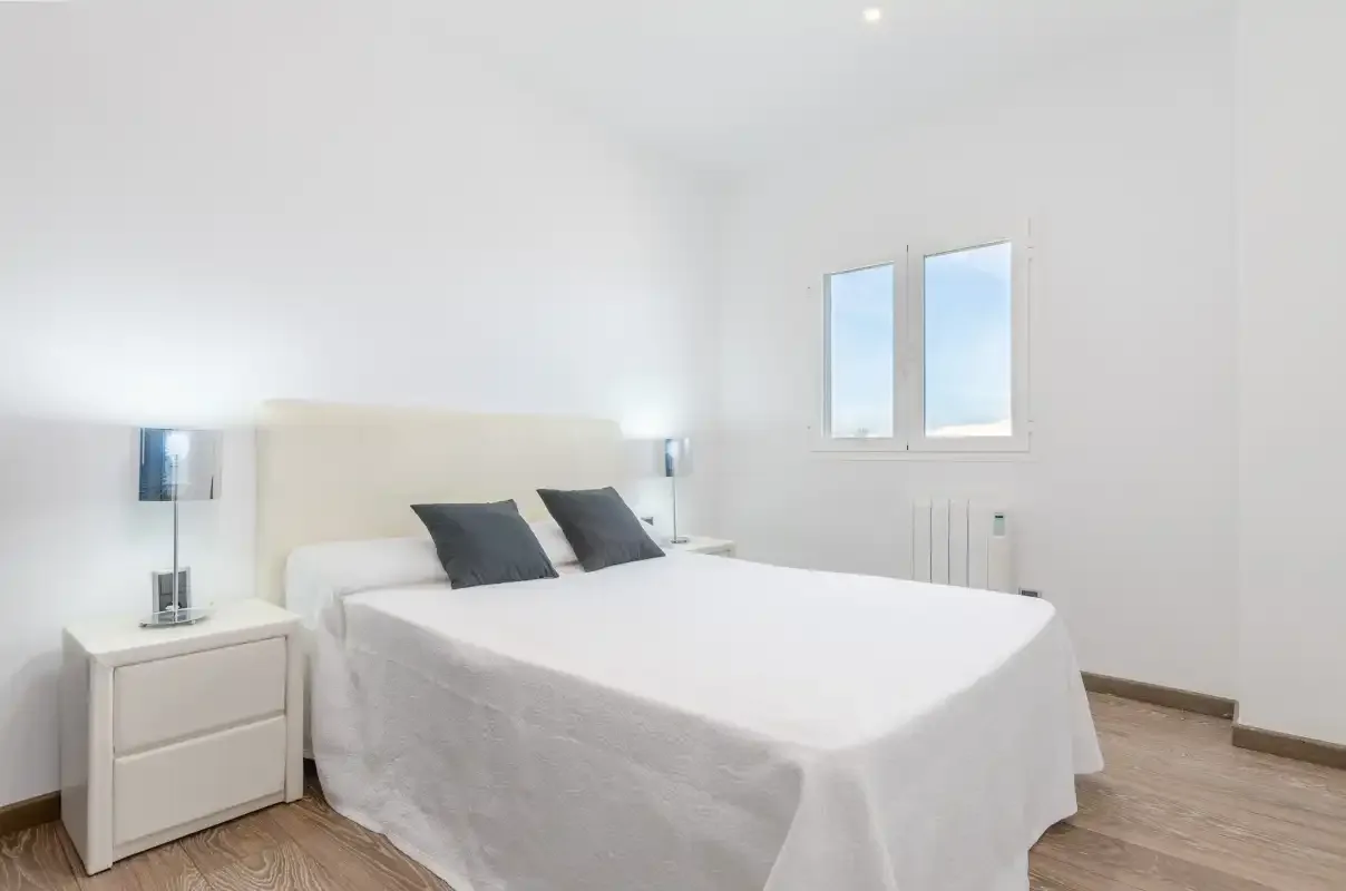 Un dormitorio luminoso con una cama blanca y dos ventanas.