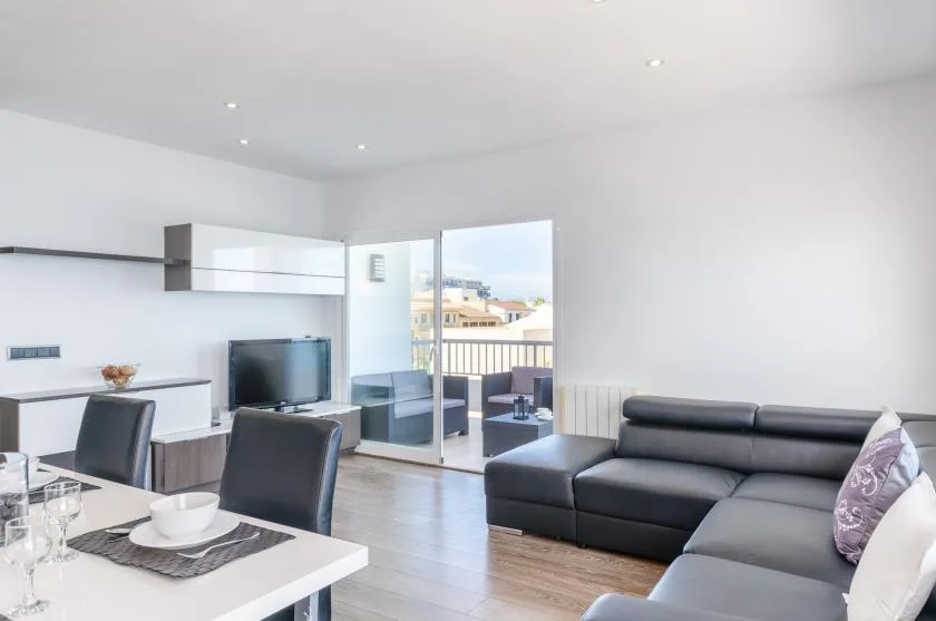 Una sala luminosa con sofá seccional y comedor junto a una puerta abierta al balcón.