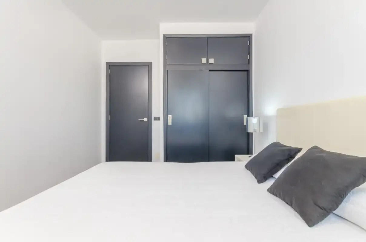 Un dormitorio con una cama blanca y almohadas oscuras junto a una puerta oscura.