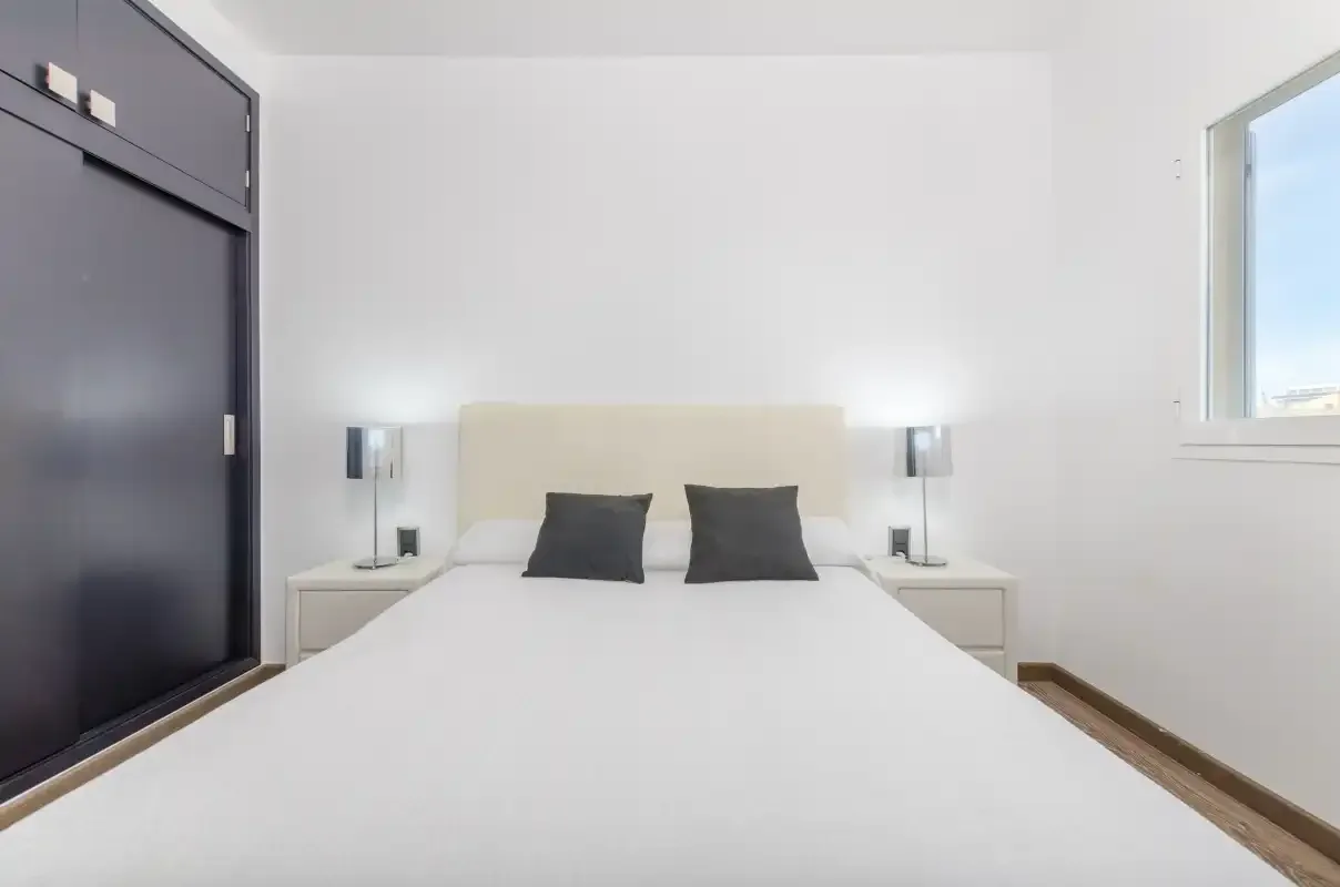 Un dormitorio luminoso con una cama blanca, almohadas oscuras y lámparas de noche.