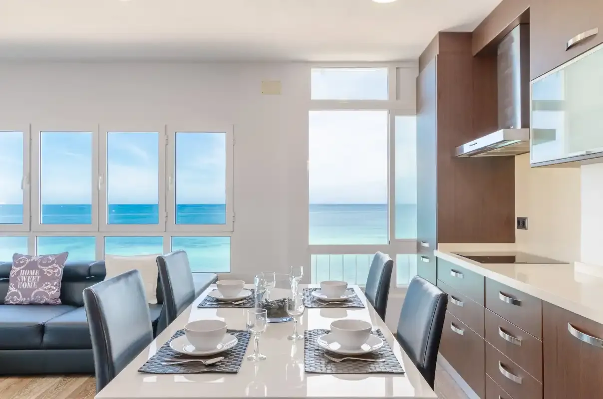 Un comedor luminoso con la mesa puesta junto a grandes ventanales con vistas al mar.