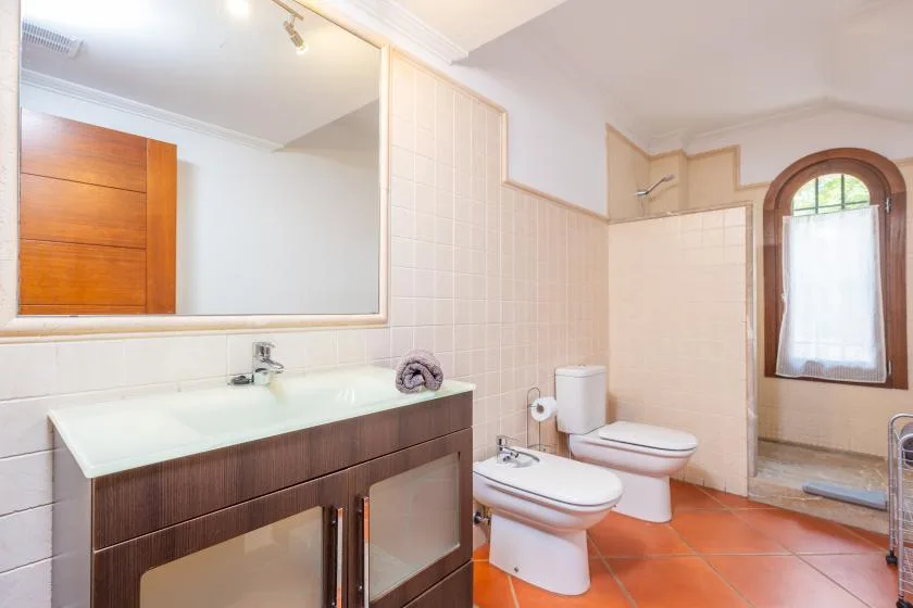 A tiled bathroom with a double sink, toilet, and arched window.