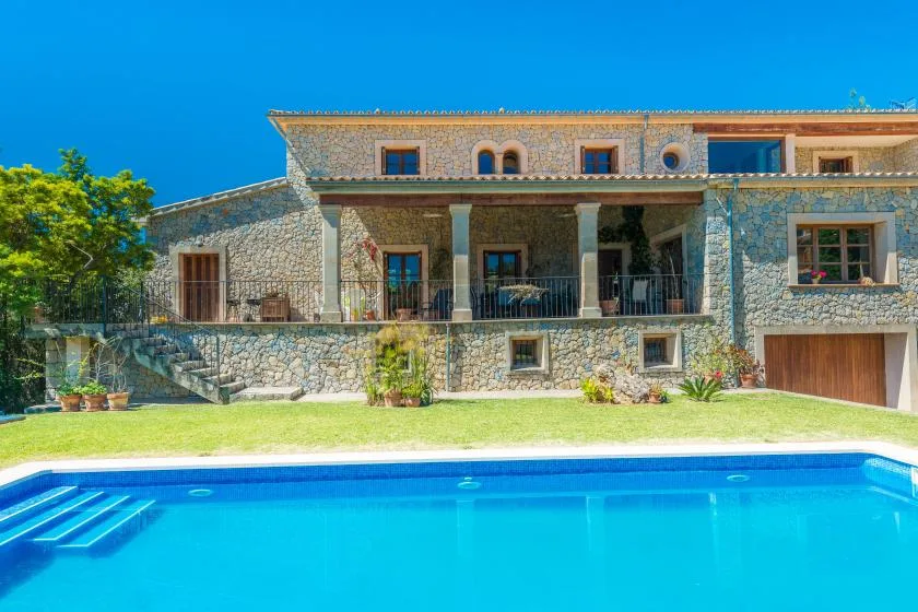 A stone house with a covered terrace overlooks a swimming pool and lawn.