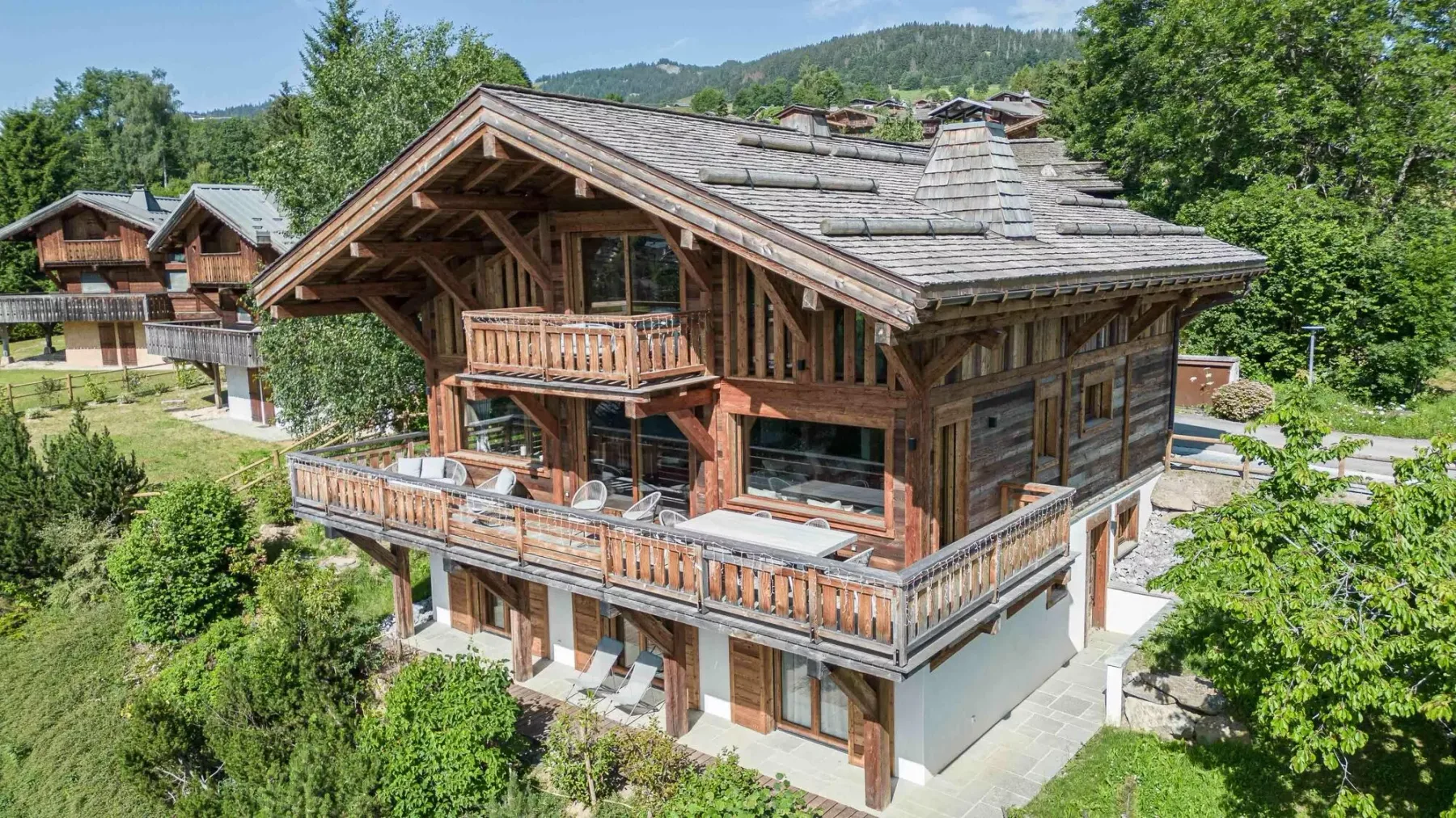 A large wooden chalet sits among green trees with forested hills in the background.