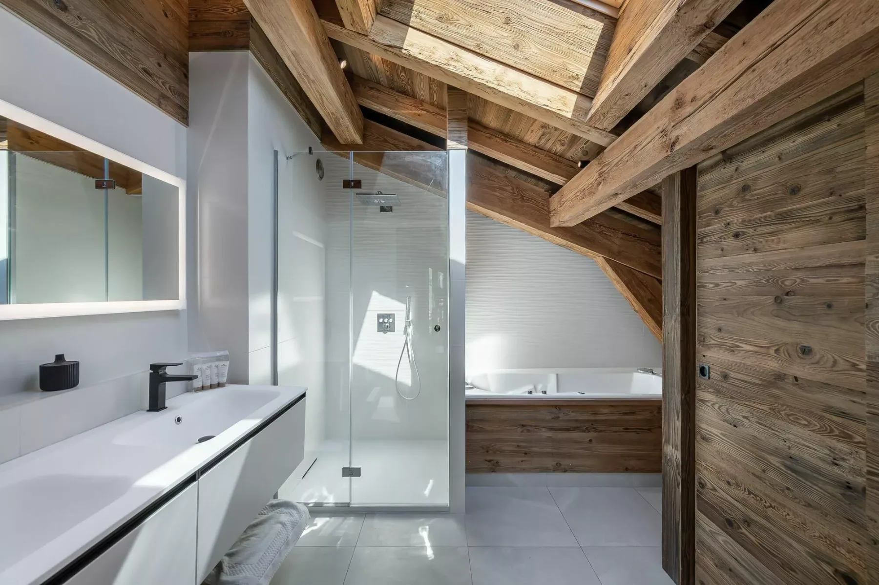 A bathroom with a glass shower and a bathtub beneath a sloped wooden ceiling.