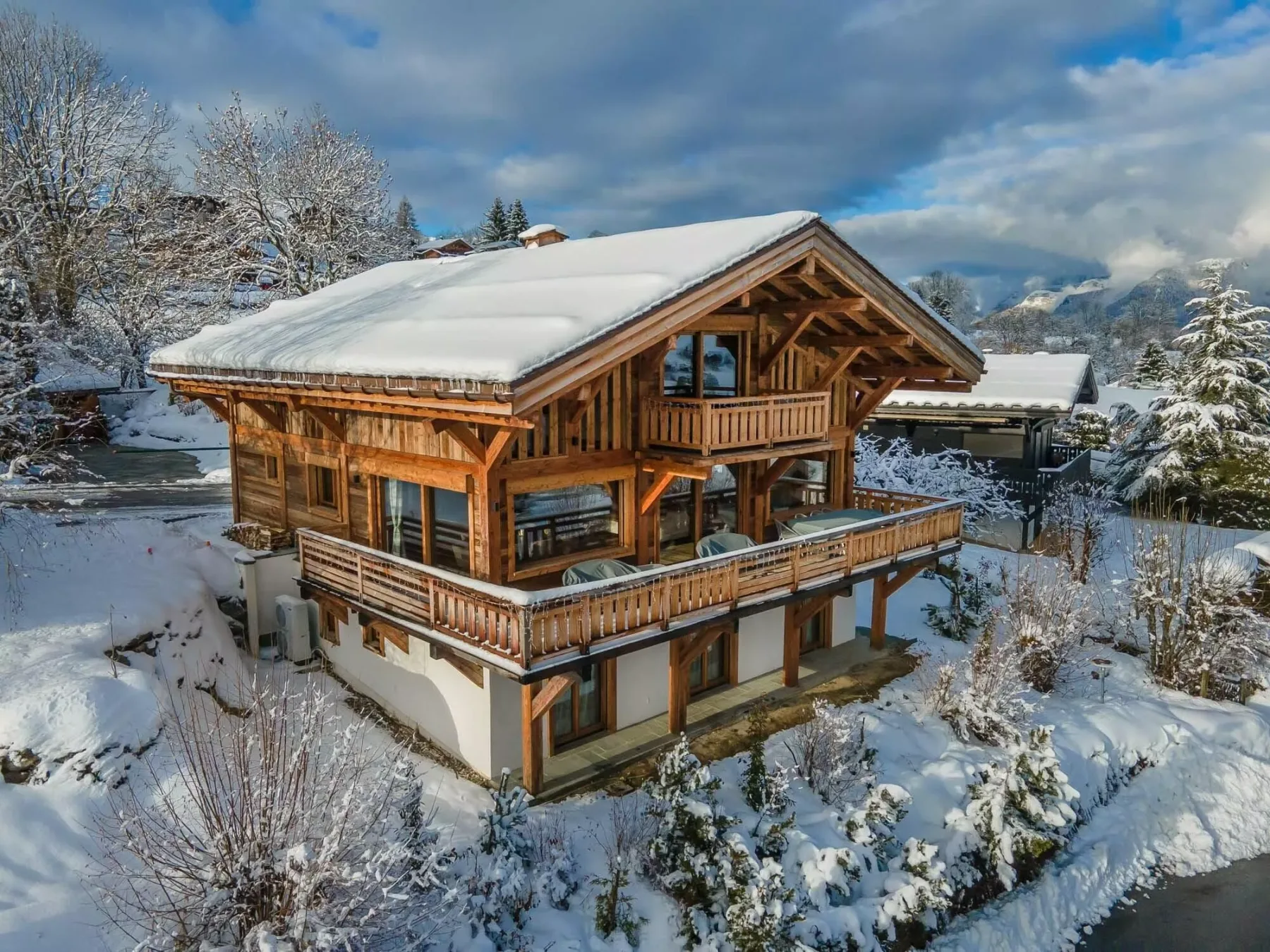 A wooden chalet stands amid snow-covered trees beneath a cloudy mountain sky.