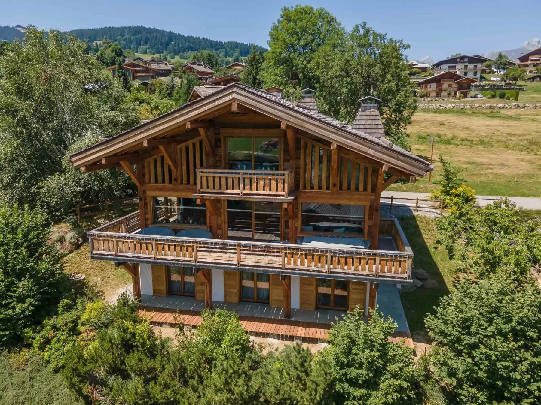 A large wooden chalet stands among trees, with hills and other homes in the background.