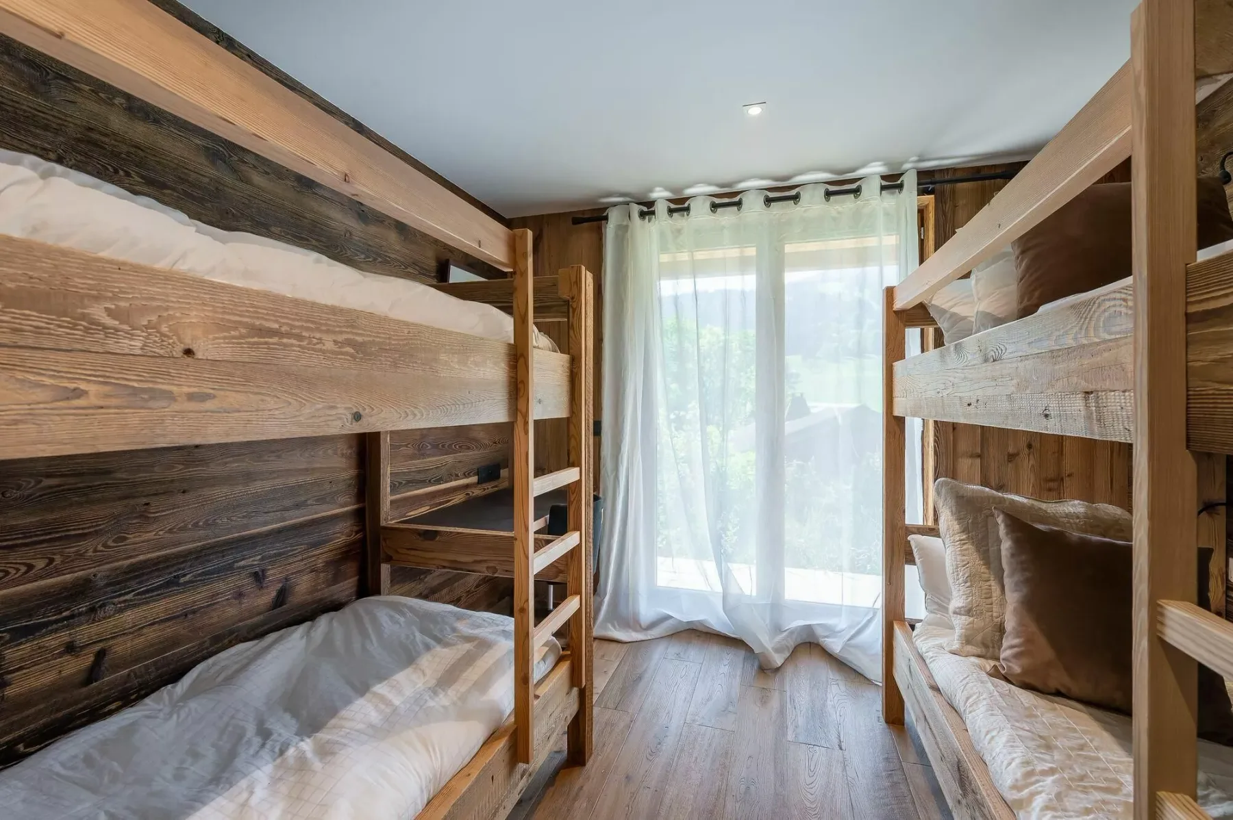 A bedroom with wooden bunk beds and a bright window behind sheer curtains.