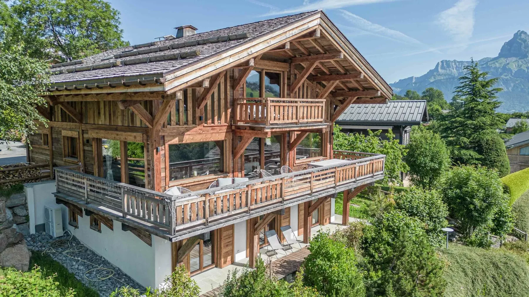 A large timber house with balconies stands among green trees beneath mountain peaks.