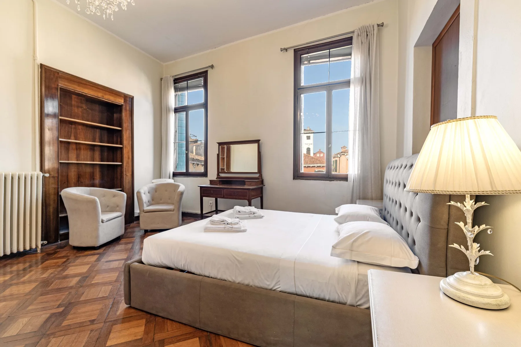A bedroom with a large bed, tall windows, and a wooden bookcase.