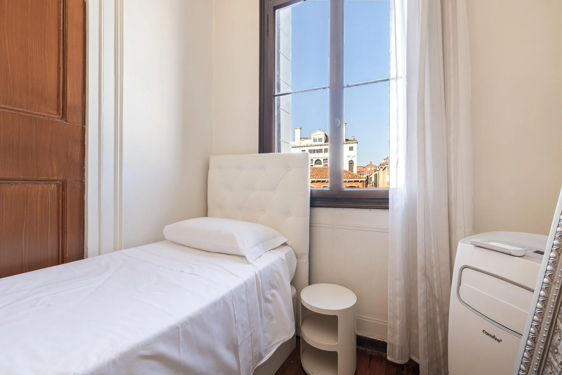 A bedroom with a single bed beside a window overlooking nearby buildings.
