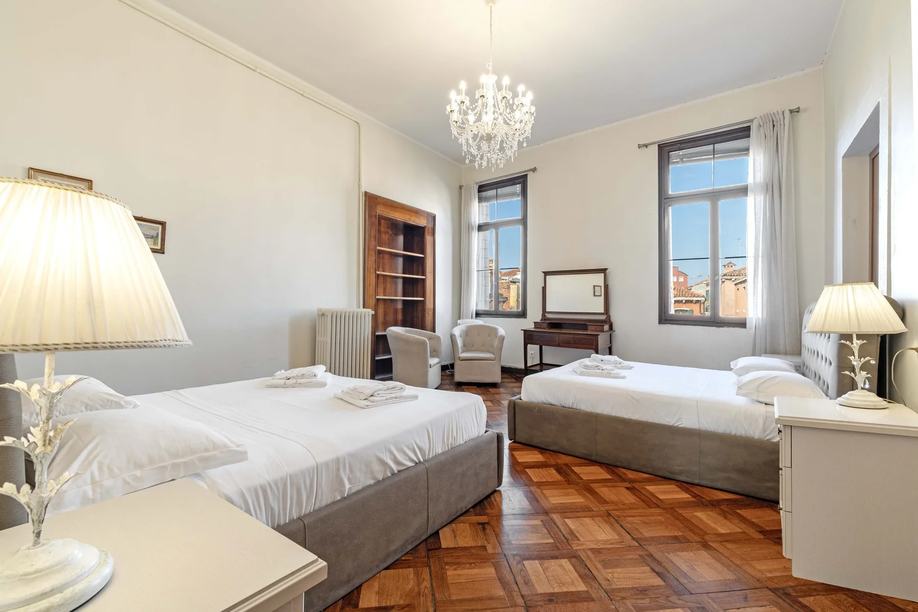 A bright bedroom with two beds, tall windows, and a chandelier.