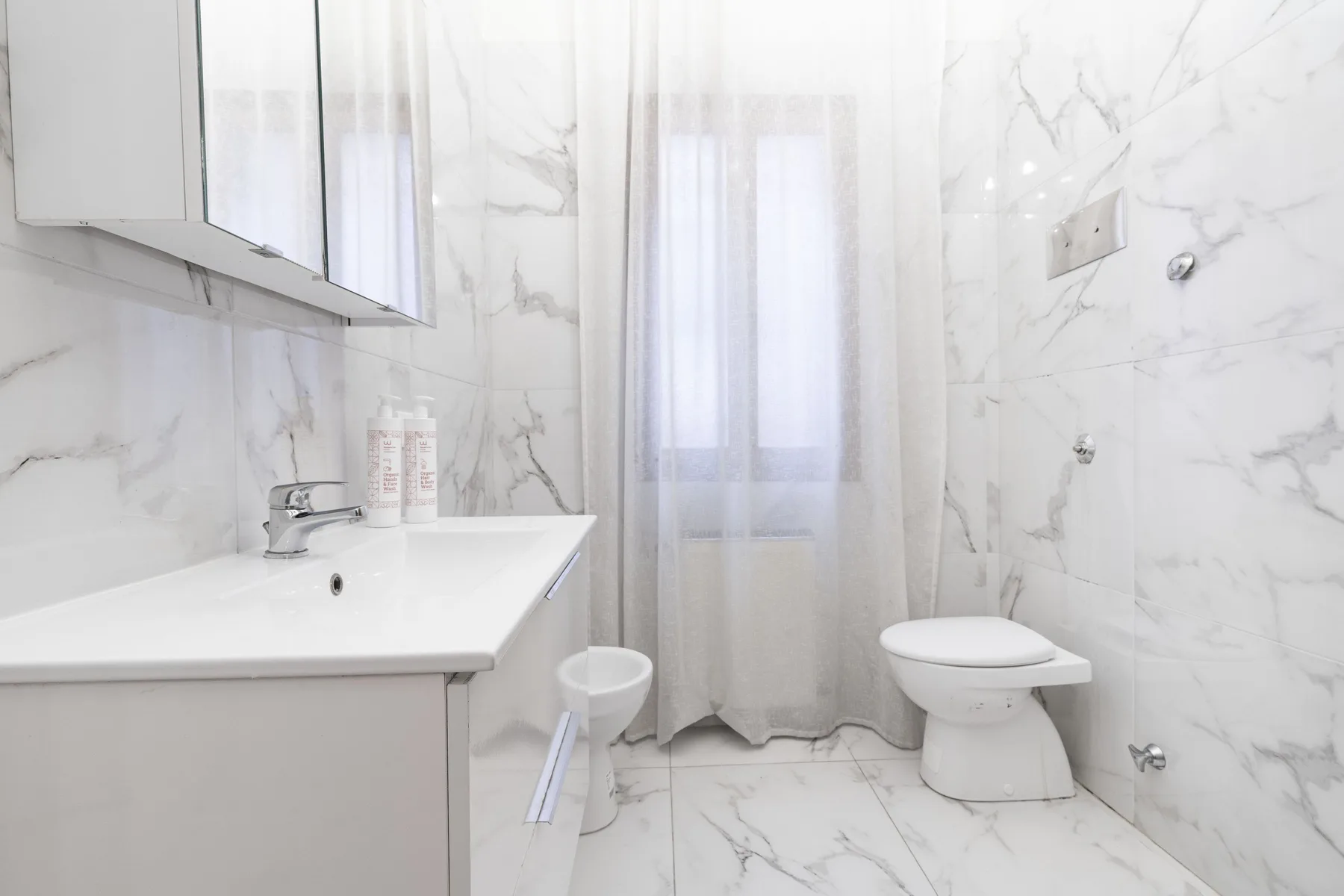 A bright bathroom with a sink, toilet, and sheer curtains over the window.