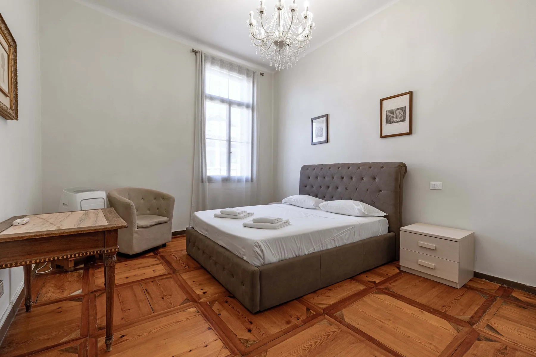 A bedroom with a bed, a small desk, and a tall curtained window.