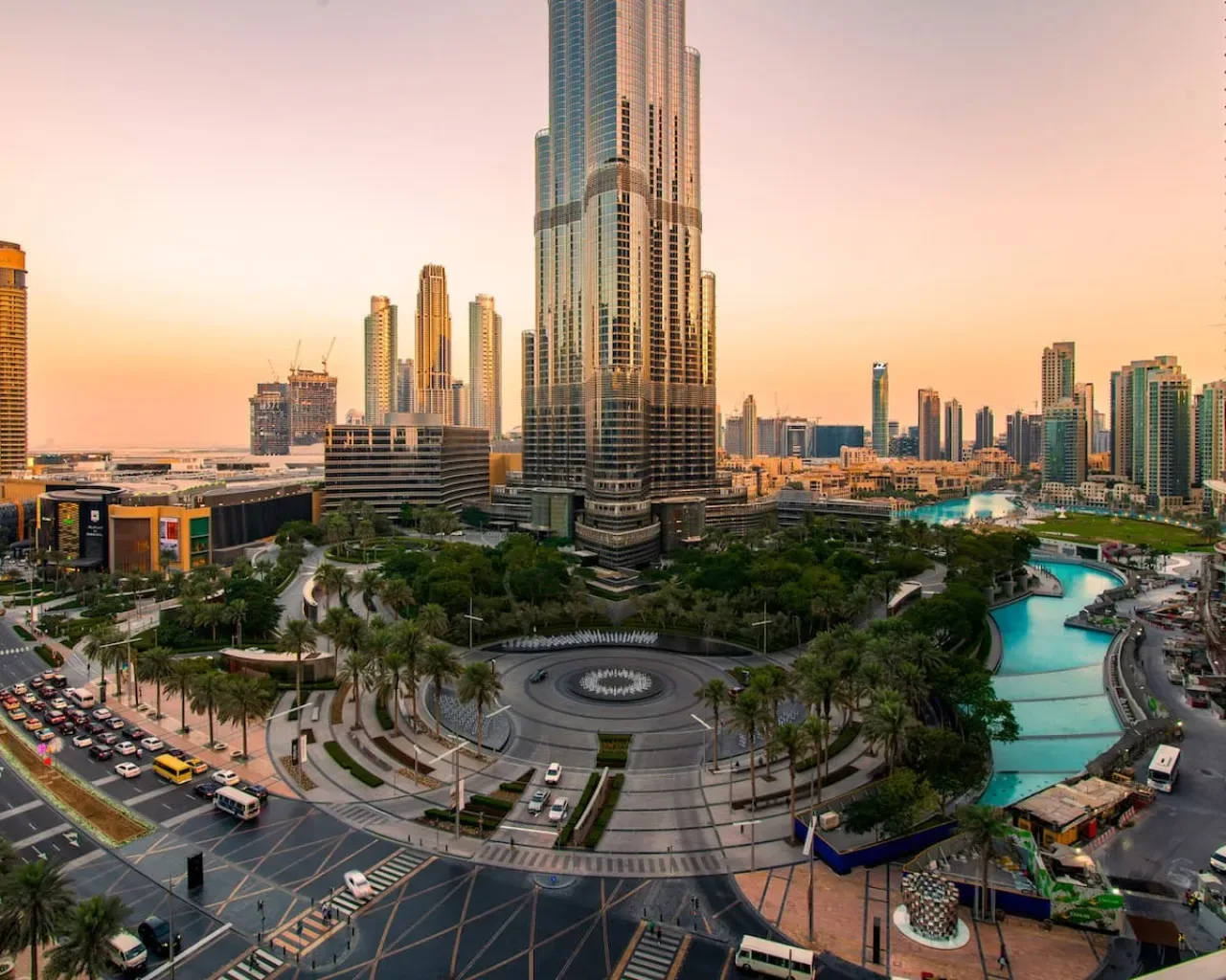 A city skyline at sunset rises above landscaped grounds and a curving turquoise waterway.