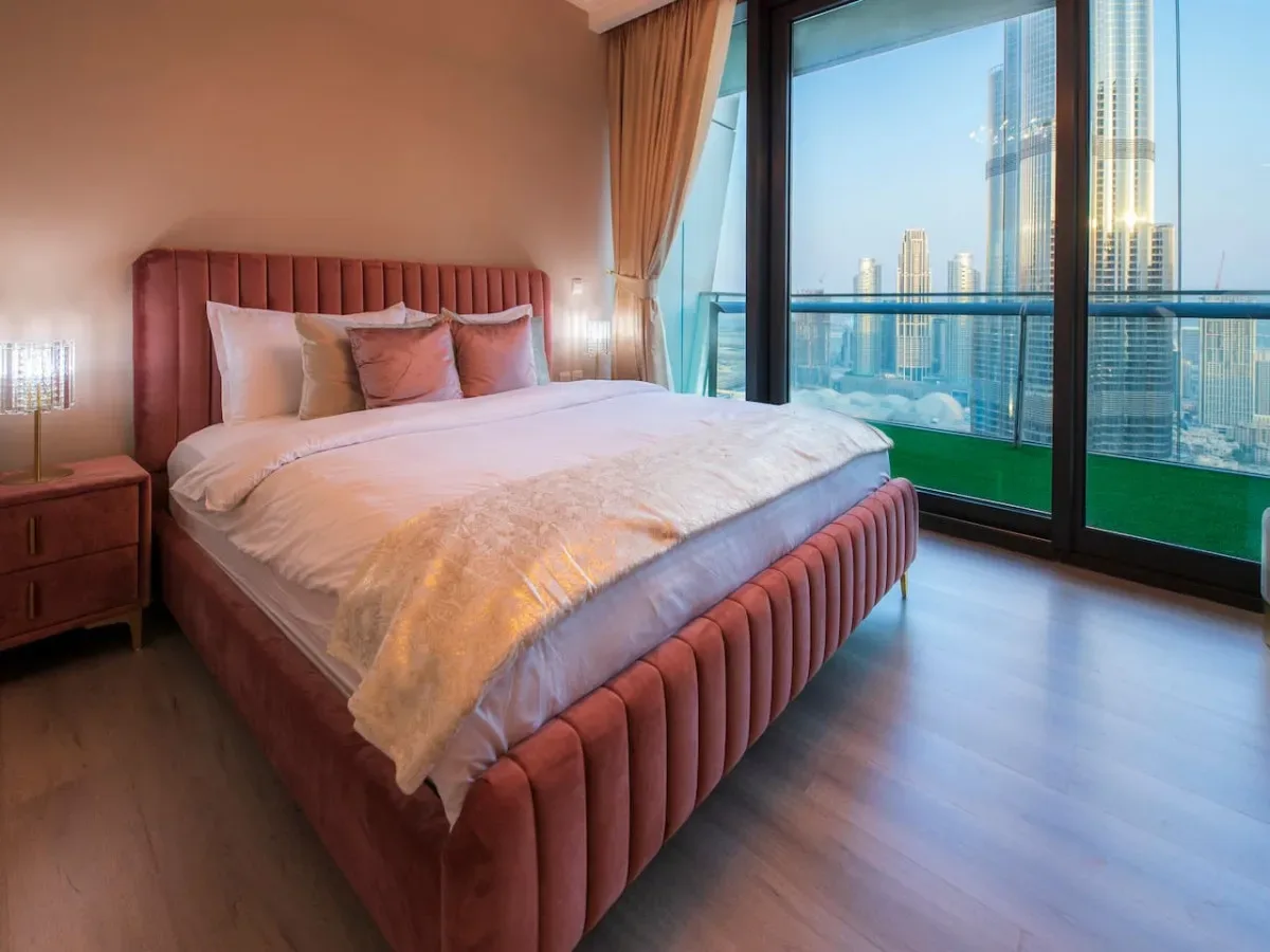 A bedroom with a neatly made bed beside floor-to-ceiling windows overlooking city buildings.