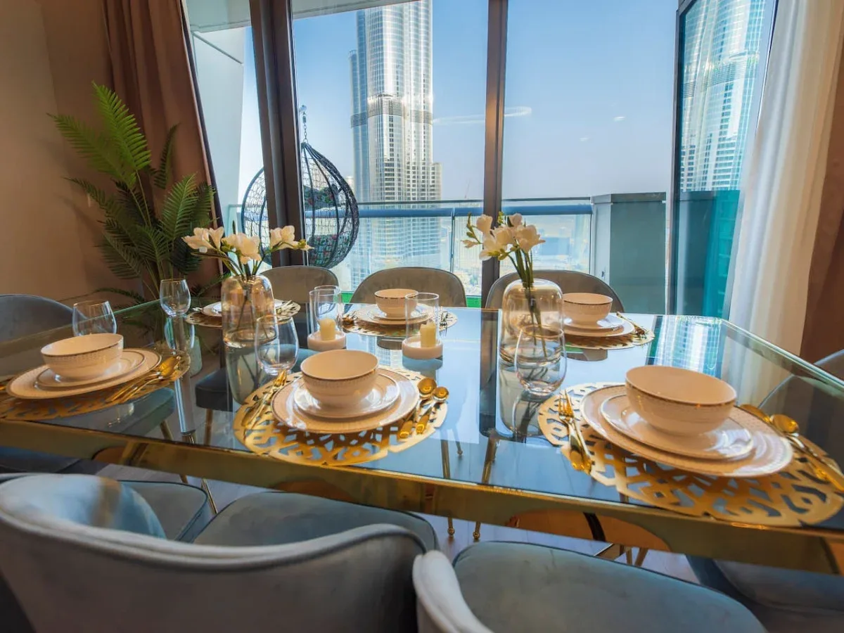 A dining table is set with place settings beside floor-to-ceiling windows overlooking nearby towers.