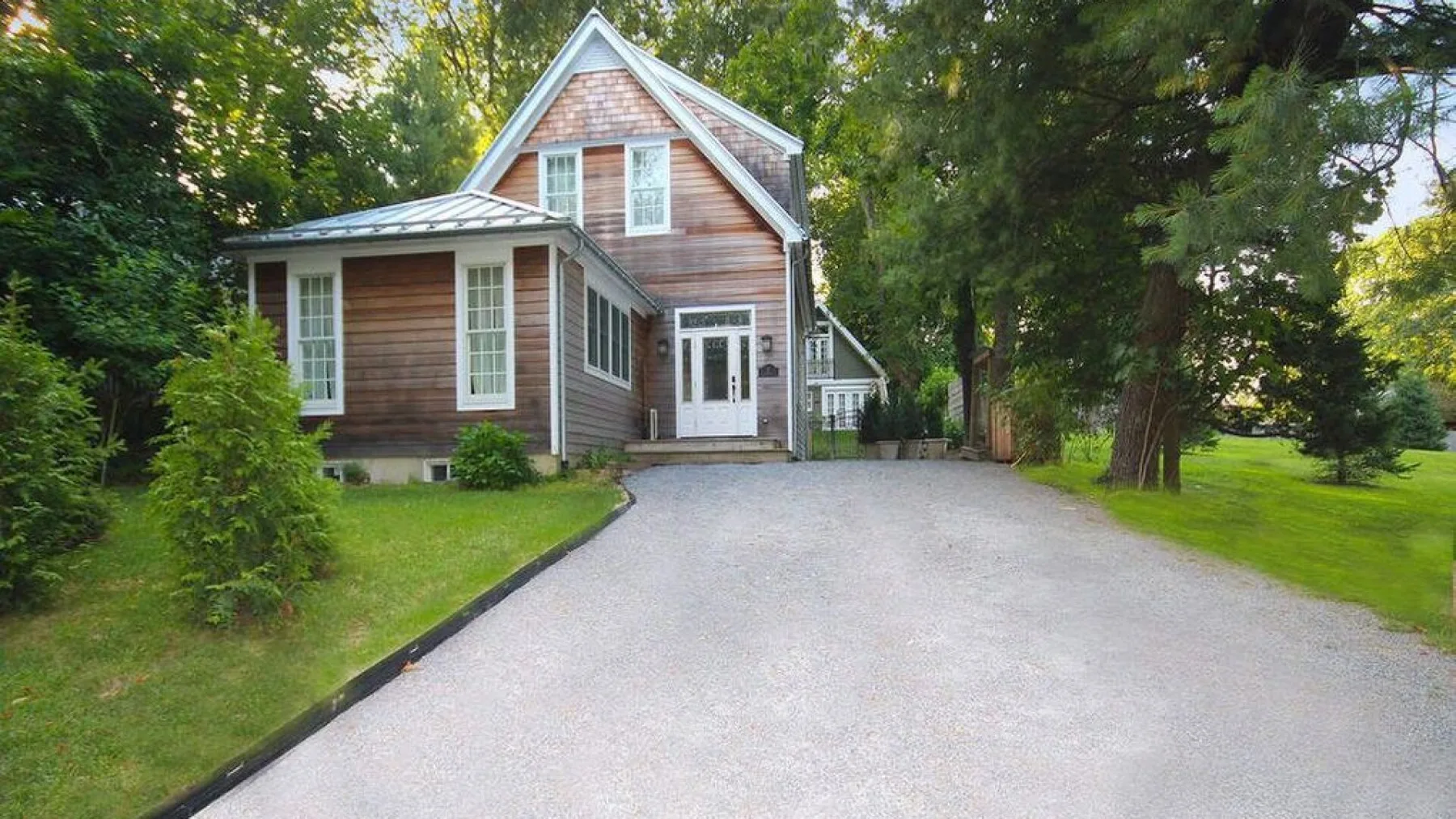 A house stands beside a paved driveway, surrounded by trees and lawn.