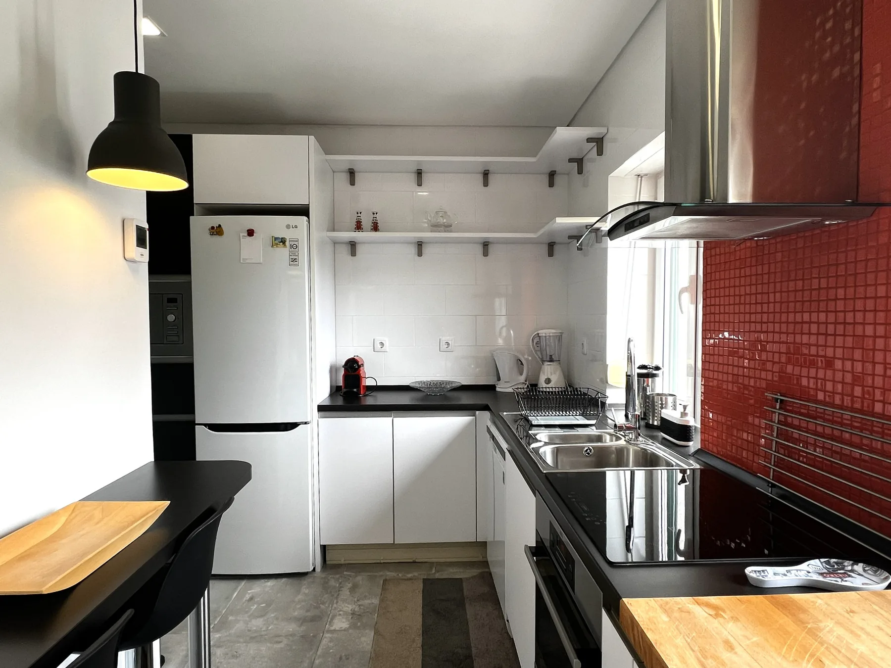 Una cocina compacta con armarios blancos, encimeras oscuras y una pared de azulejos rojos.