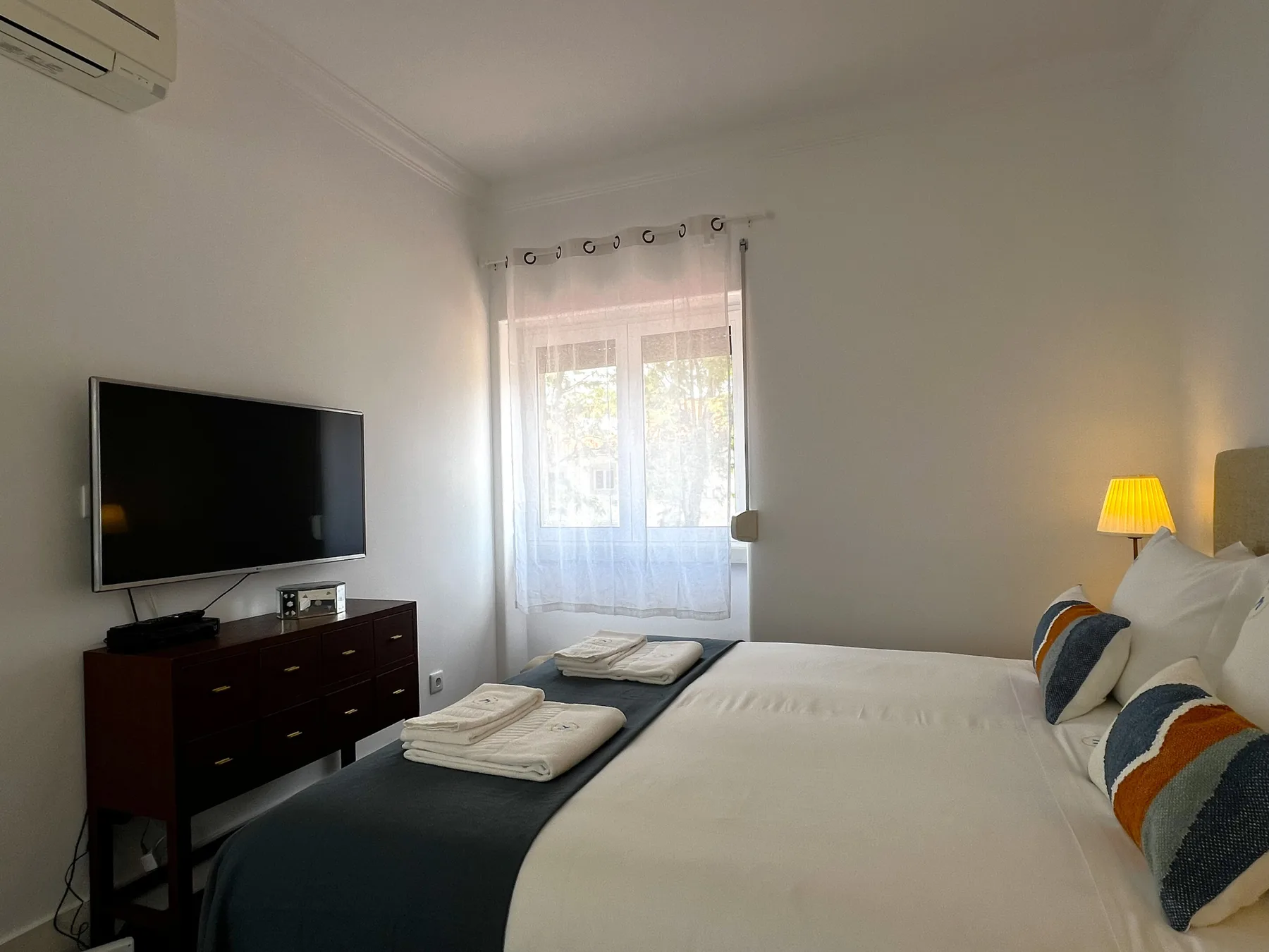 Un dormitorio luminoso con una cama grande, un televisor y una ventana con cortinas transparentes.