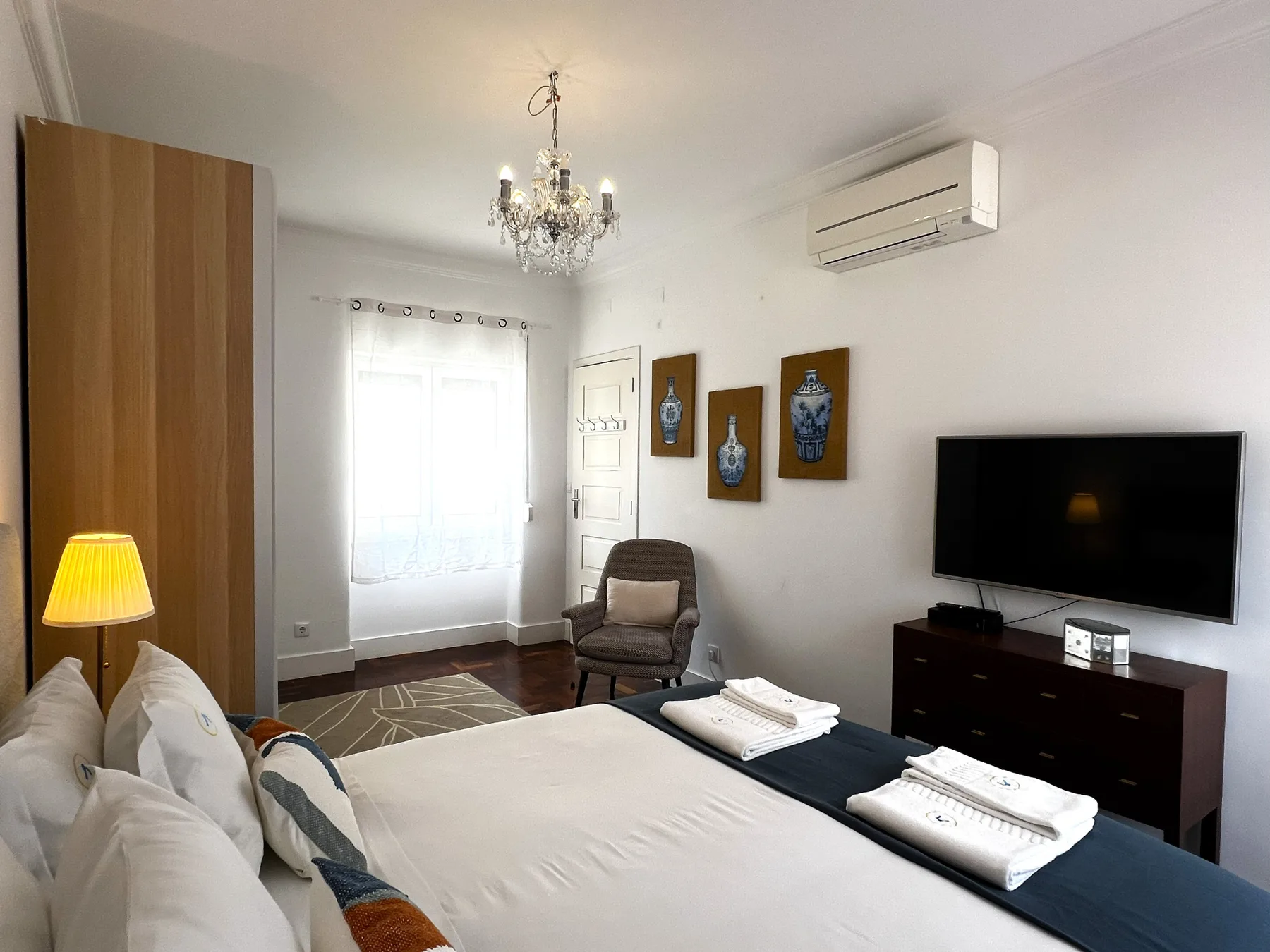 Un dormitorio con una cama bien hecha, un televisor y un sillón junto a la ventana.