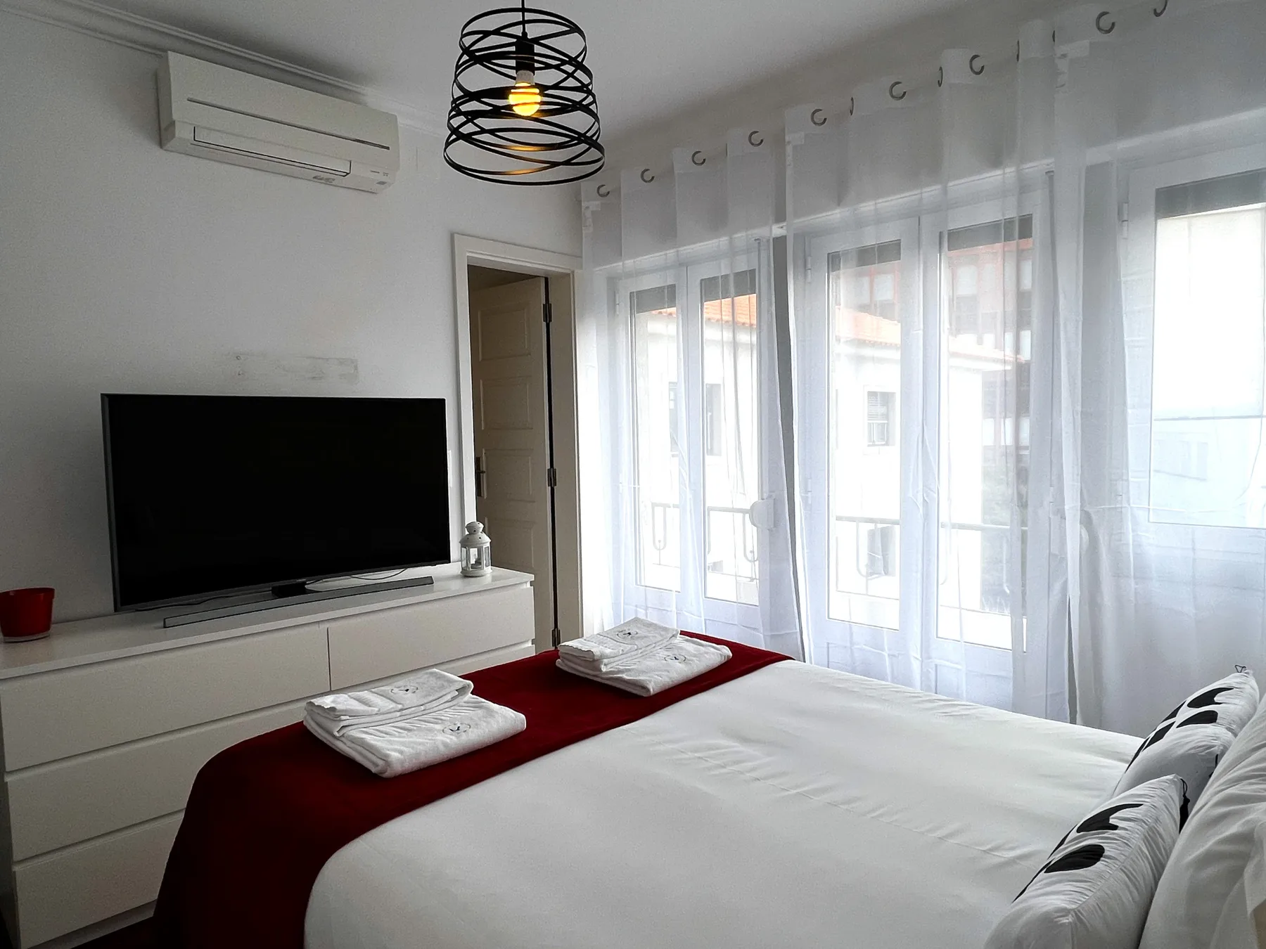 Un dormitorio luminoso tiene una cama bien tendida junto a cortinas translúcidas y ventanas grandes.