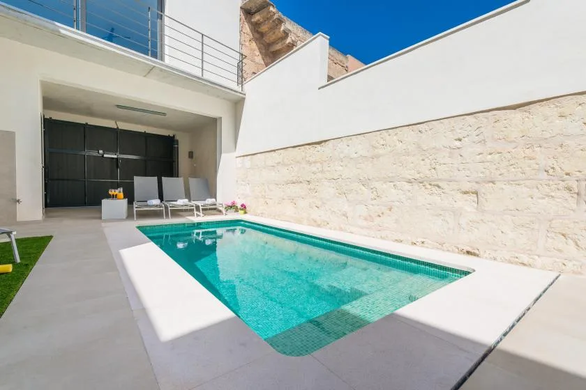 A rectangular swimming pool sits in a sunlit outdoor courtyard beside a pale stone wall.