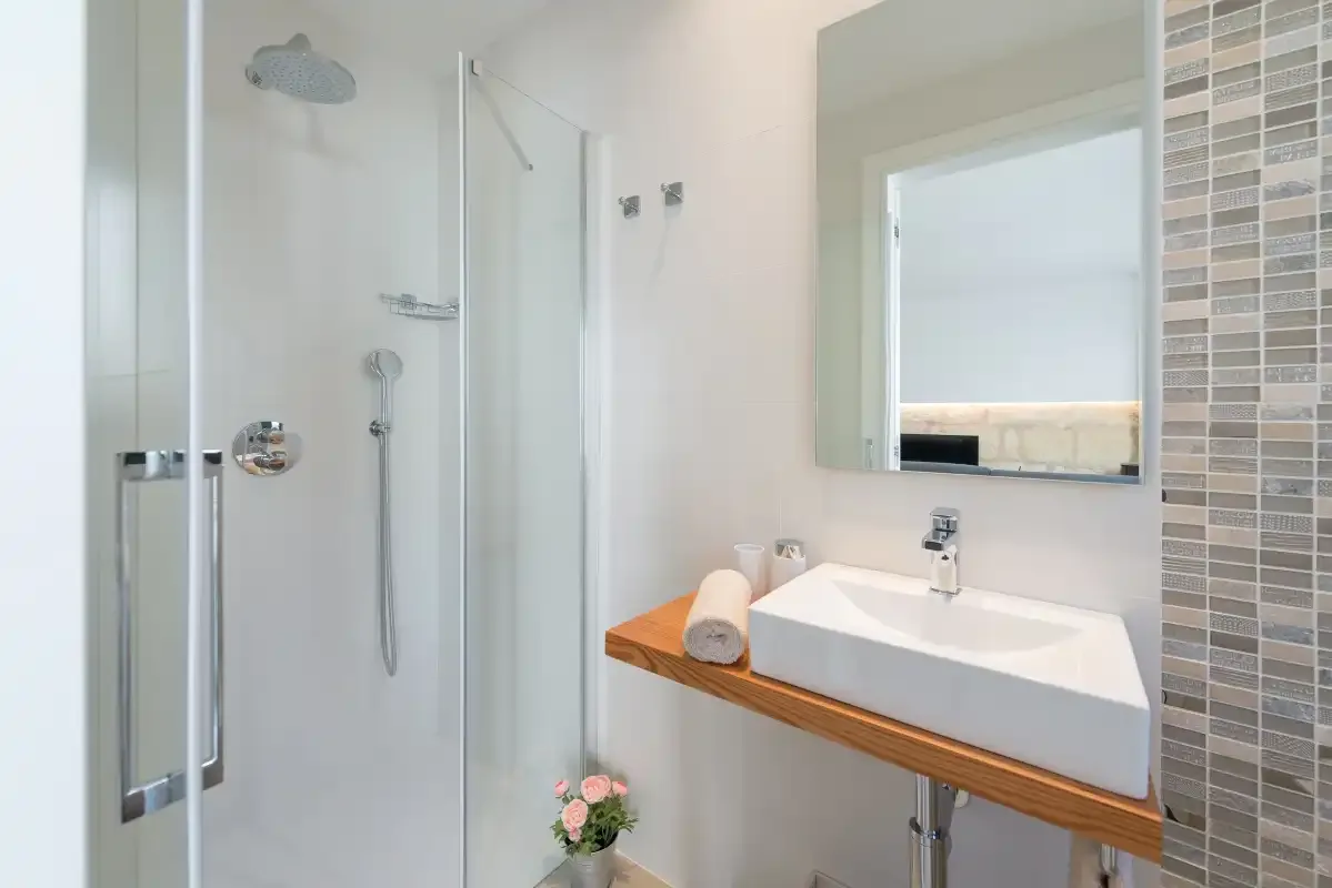 A bright bathroom has a glass shower and a white sink on a wooden counter.