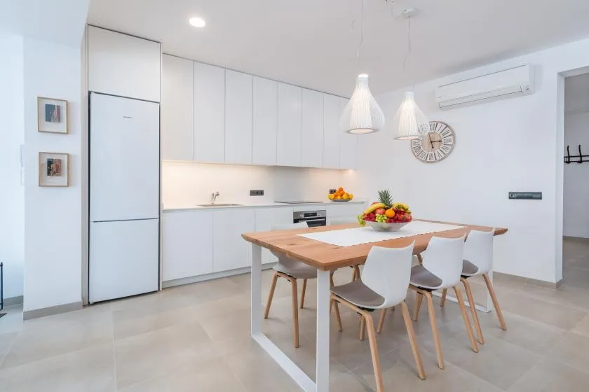 A bright dining area with a wooden table and white seating beside a modern kitchen.