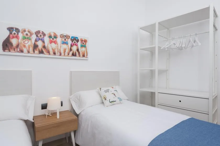 A bright bedroom with white bedding, bedside furniture, and open wardrobe shelving.