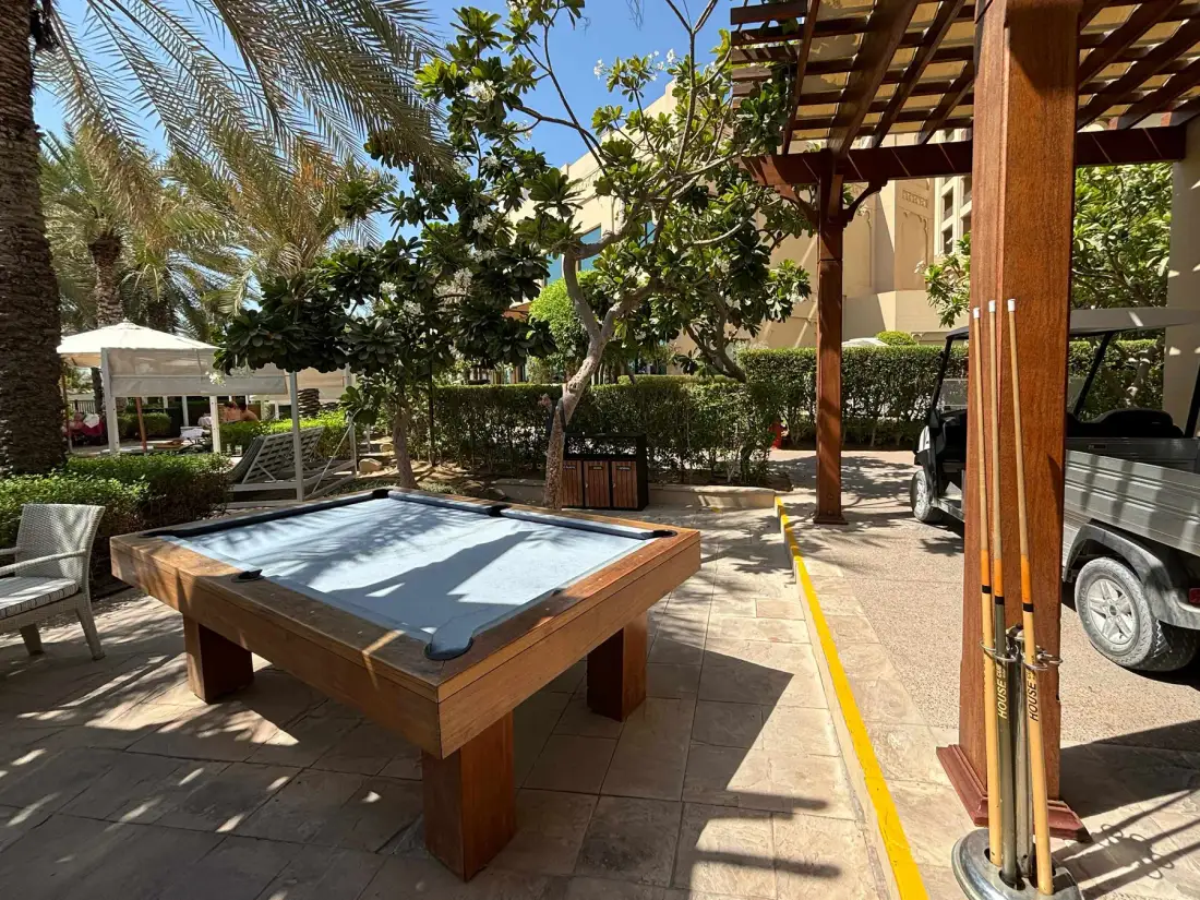 Una mesa de billar al aire libre está en un patio pavimentado bajo una pérgola de madera.