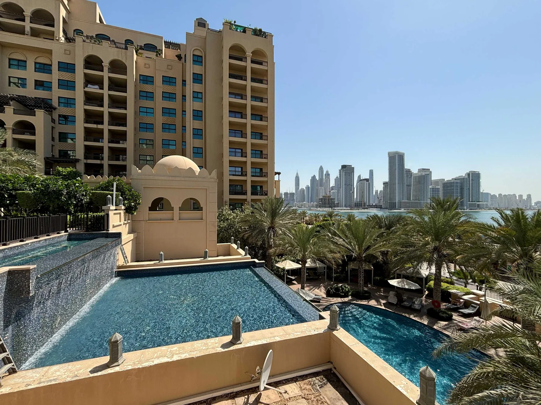 Una piscina junto a un edificio alto, con palmeras y un horizonte urbano a lo lejos.