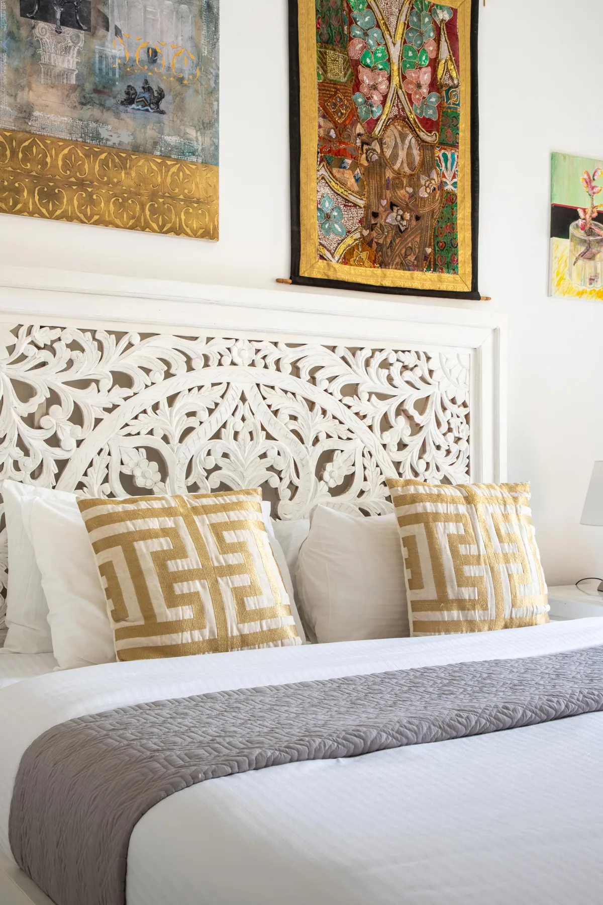 Un dormitorio con ropa de cama blanca, cojines estampados y decoración tallada sobre la cama.