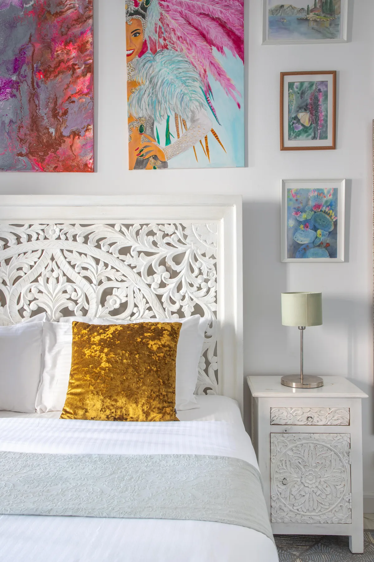 Un dormitorio con una cama blanca, un cabecero ornamentado y obras de arte coloridas en la pared.