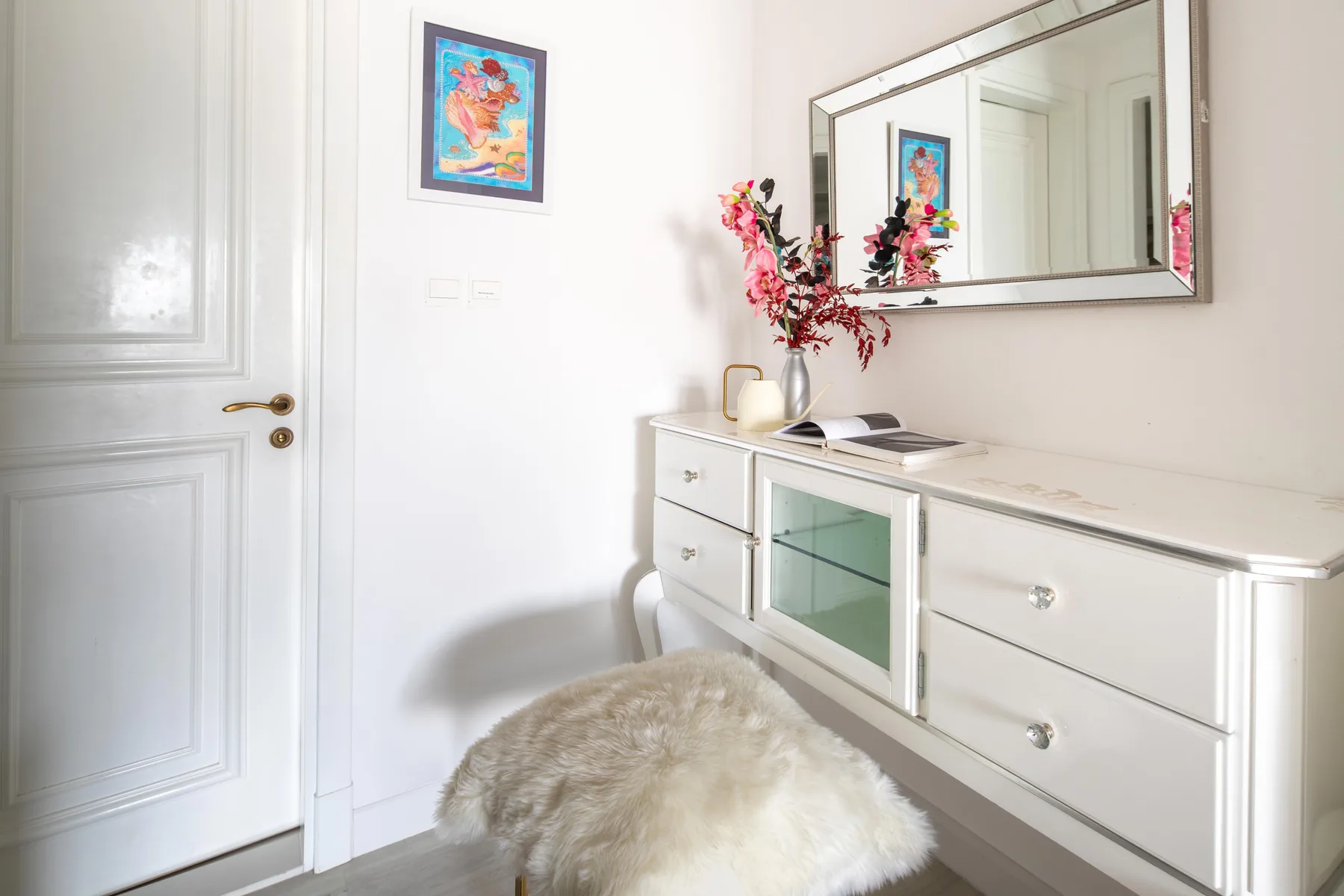 Un rincón luminoso de dormitorio con cómoda blanca, espejo de pared y cuadro enmarcado.