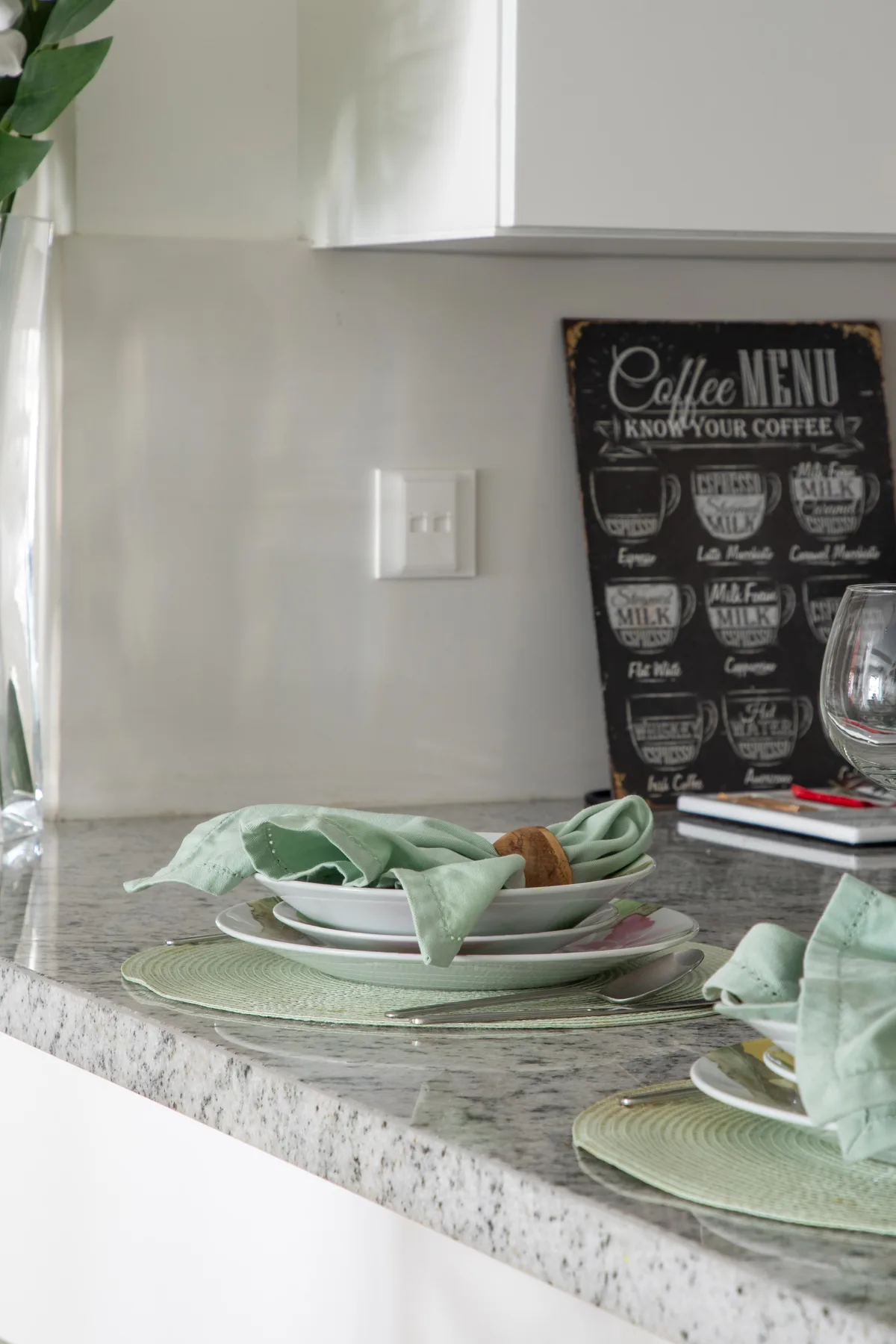 Una encimera de cocina muestra platos apilados, servilletas verdes y un cartel de café.