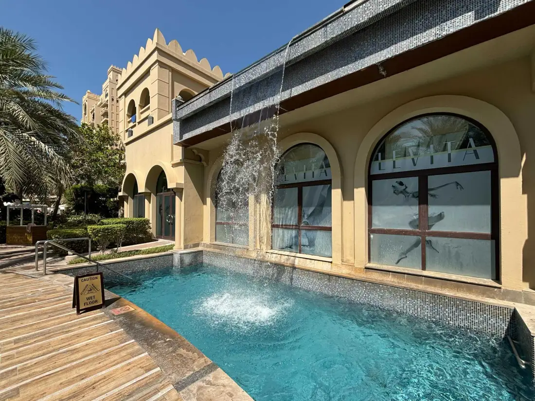 Una piscina de patio está junto a una fachada con arcos, con agua cayendo en ella.